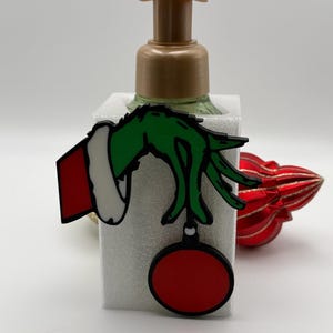 May include: A rectangular soap dispenser with a tan pump mechanism features a decorative attachment of a green hand with a white-trimmed red sleeve holding a red circular ornament. The attachment is secured to the front of the white container.