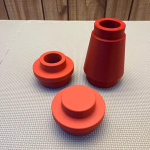 May include: Three red, geometric, 3D-printed objects. One is a truncated cone, another is a short cylinder, and the third is a stepped cylinder. The objects are arranged on a light gray surface with a wood-paneled background.