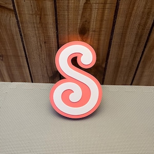 May include: A layered, coral-colored wooden letter "S" with a white, swirled design. The decorative initial stands on a light gray surface against a wooden background.