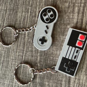 Peut inclure: Deux porte-clés représentant des manettes de jeu vidéo classiques. L'un est une manette grise avec des boutons noirs et une croix directionnelle en forme de plus. L'autre est une manette noire avec des boutons rouges et une croix directionnelle en forme de plus.