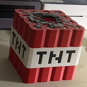 Könnte beinhalten: Ein rot-weißer TNT-Block mit dem Wort "TNT" auf der Seite. Die Oberseite des Blocks hat ein graues, pixeliges Design. Bei diesem Artikel handelt es sich wahrscheinlich um ein Dekorationsobjekt oder ein Spielzeug.