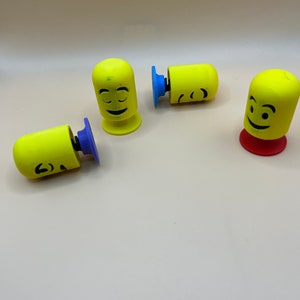 May include: Four yellow phone holders with different facial expressions. Each holder has a round base with a suction cup on the bottom. The suction cups are blue, purple, and red.