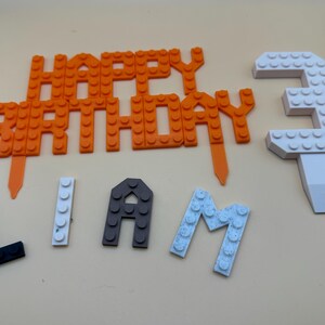 3D printed LEGO-style brick cake topper set for a 3rd birthday, featuring an orange "Happy Birthday" sign, a large white number 3, and personalized name letters for "Liam" in black, white, and grey.