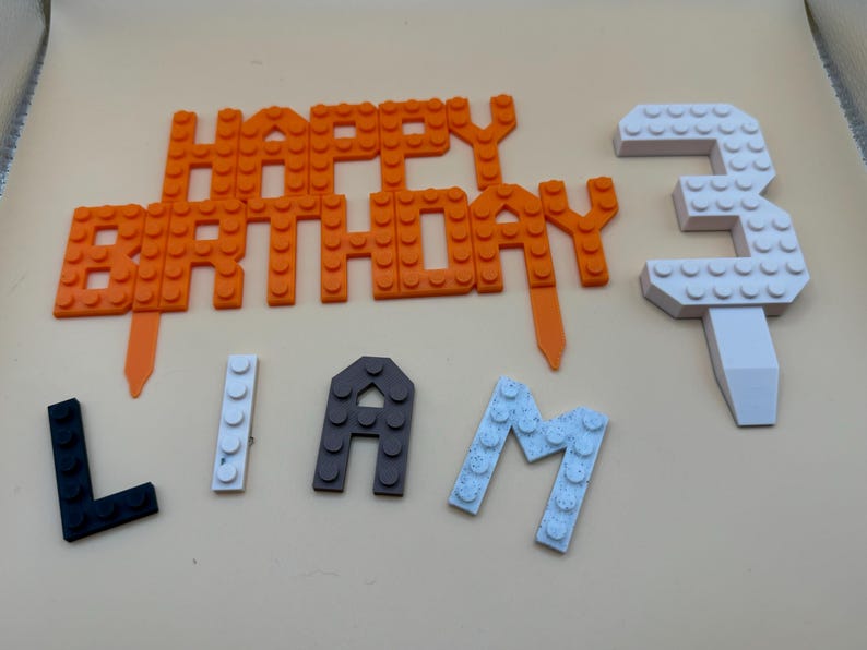 3D printed LEGO-style brick cake topper set for a 3rd birthday, featuring an orange "Happy Birthday" sign, a large white number 3, and personalized name letters for "Liam" in black, white, and grey.