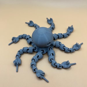May include: A gray 3D printed octopus with eight articulated arms, each ending in a hand with a pointing finger. The octopus is posed with all arms pointing in different directions.