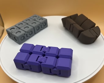 Puede incluir: Tres juguetes antiestrés, uno es un cubo morado, otro es un cubo gris y el último es un cubo marrón.