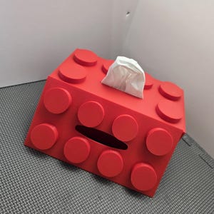 May include: Red plastic tissue box shaped like a Lego brick. The box has a slot for pulling out tissues.