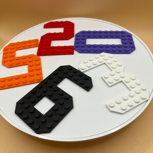 Puede incluir: Una base circular blanca con cinco números coloridos hechos de bloques de Lego. Los números son 5, 2, 0, 6 y 3. Los números están dispuestos en un círculo alrededor de la base.