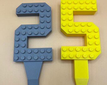 May include: Two number-shaped cake toppers. One is a gray number 2, and the other is a yellow number 5. Both are designed to resemble building blocks and have a stake at the bottom for insertion into a cake or other dessert.
