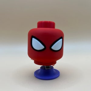 May include: A red and white Lego-style figure of Spider-Man's head with a blue base. The figure is a toy.