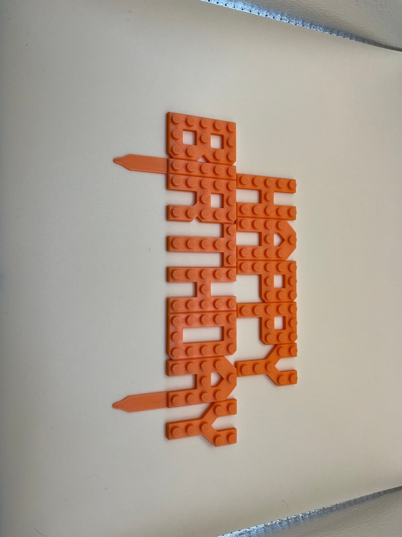 Orange 3D-printed plastic 'Happy Birthday' cake topper with a building-block brick texture design.