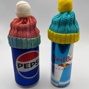 Könnte beinhalten: Zwei Getränkedosen, eine Pepsi und eine Red Bull, jeweils mit einer kleinen Strickmütze versehen. Die Pepsi-Dose hat eine rot-rosa-blaue Mütze mit einem weißen Bommel. Die Red Bull-Dose hat eine blau-gelbe Mütze mit einem gelben Bommel.