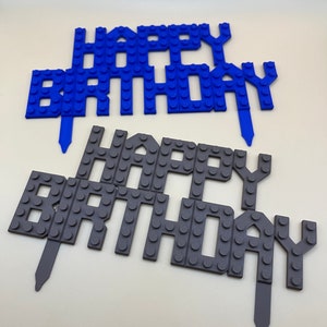 Peut inclure: Deux d&eacute;corations de g&acirc;teau en plastique bleu et gris qui disent "Joyeux anniversaire". Les d&eacute;corations sont faites pour ressembler &agrave; des briques Lego.
