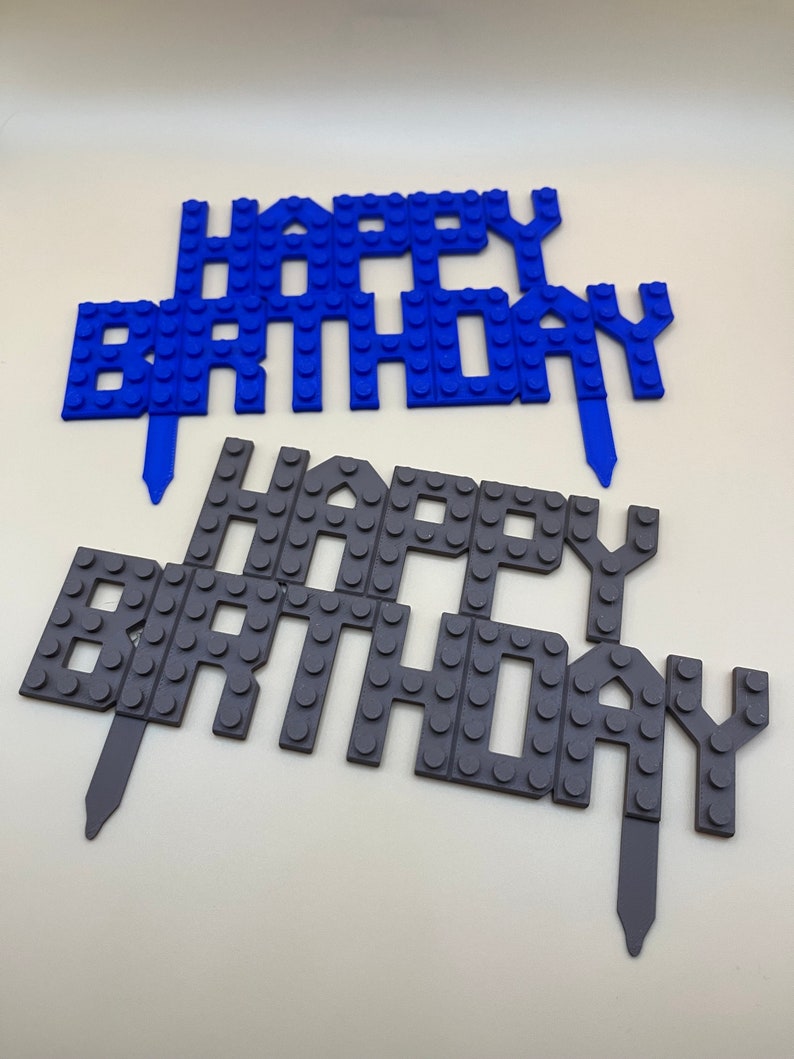 Peut inclure: Deux d&eacute;corations de g&acirc;teau en plastique bleu et gris qui disent "Joyeux anniversaire". Les d&eacute;corations sont faites pour ressembler &agrave; des briques Lego.