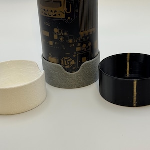 Może przedstawiać: Czarny i złoty cylindryczny pojemnik z wzorem płytki drukowanej, spoczywający w szarej podstawce, w towarzystwie białego i czarnego okrągłego uchwytu. Przedmioty znajdują się na białej powierzchni.