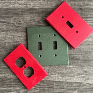 May include: Three painted wall plates, one with a double light switch, one with a single light switch, and one with a double outlet. The plates are painted in red and green.