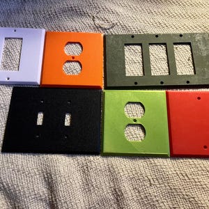 May include: A selection of colourful electrical outlet and switch plates. The plates are in various colours, including purple, orange, black, green, and red. The plates have different configurations for outlets and switches.