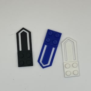 May include: Three plastic bookmark clips in black, blue, and white. Each clip has a rectangular shape with a small, raised section of three circles at the bottom. The clips are designed to look like Lego bricks.