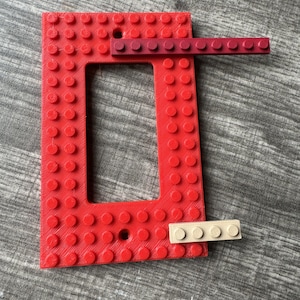 May include: A red plastic light switch cover shaped like a Lego brick. The cover has a rectangular opening for the switch. There are two small Lego bricks attached to the cover, one red and one beige.