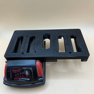 Pode incluir: Um suporte de bateria de plástico preto com ranhuras para várias baterias. Uma bateria Milwaukee REDLITHIUM vermelha e preta está no suporte.