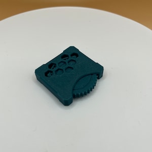 May include: A dark teal plastic gear with a honeycomb pattern on the top. The gear is designed to be used with a 3D printer.