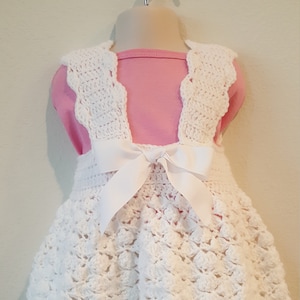 May include: A white crochet dress with a matching top and a large white bow at the back. The dress is displayed on a mannequin and is likely for a baby or toddler. The dress has a textured, open weave design.