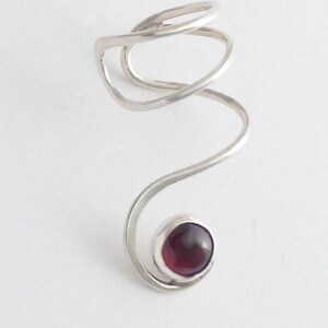 May include: A silver-toned metal ring featuring a spiraling, open-ended band design. The structure curves upward into a circular loop at the top and terminates at the bottom with a single, smooth, dark red cabochon stone set in a silver bezel.