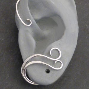 May include: A silver ear cuff with a curved design, featuring a spiral at the top and a double-loop design. The jewelry is displayed on a gray ear model, showcasing the intricate details of the metalwork.