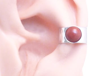May include: A silver-toned ear cuff featuring a wide, hammered metal band with a smooth, circular, reddish-brown stone set in the center. The jewelry piece is positioned on the outer cartilage of an ear, showcasing a polished, reflective finish.
