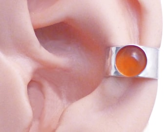 May include: A silver ear cuff featuring a smooth, wide band with a hammered texture and a single, circular, polished orange carnelian gemstone set flush into the metal. The piece is designed to wrap around the outer edge of the ear cartilage.