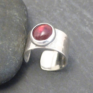 May include: A silver ring with a red gemstone set in a bezel setting. The ring is wide and has a hammered texture.