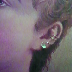 Pode incluir: Um ear cuff de tom prateado com um design de fio curvo e liso que envolve a cartilagem da orelha, acentuado com uma &uacute;nica pedra redonda pequena de caboch&atilde;o verde-p&aacute;lido fixada na base do l&oacute;bulo da orelha para um visual minimalista.