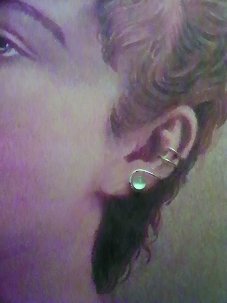 Pode incluir: Um ear cuff de tom prateado com um design de fio curvo e liso que envolve a cartilagem da orelha, acentuado com uma &uacute;nica pedra redonda pequena de caboch&atilde;o verde-p&aacute;lido fixada na base do l&oacute;bulo da orelha para um visual minimalista.