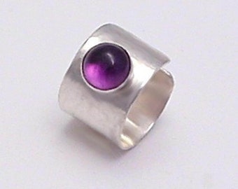 May include: A wide silver band ring featuring a single, circular, polished purple amethyst gemstone set into the metal surface. The ring has a smooth, reflective finish and a consistent width throughout its circular structure.