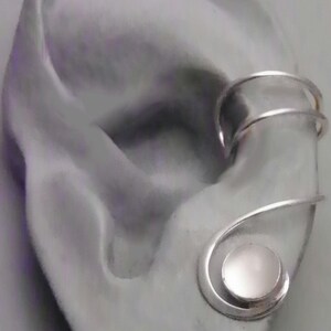 Pode incluir: Um ear cuff de tom prateado com uma banda de fio duplo que envolve a cartilagem superior da orelha e termina numa pedra caboch&atilde;o circular de cor clara, engastada numa estrutura de metal em espiral na parte inferior da orelha.