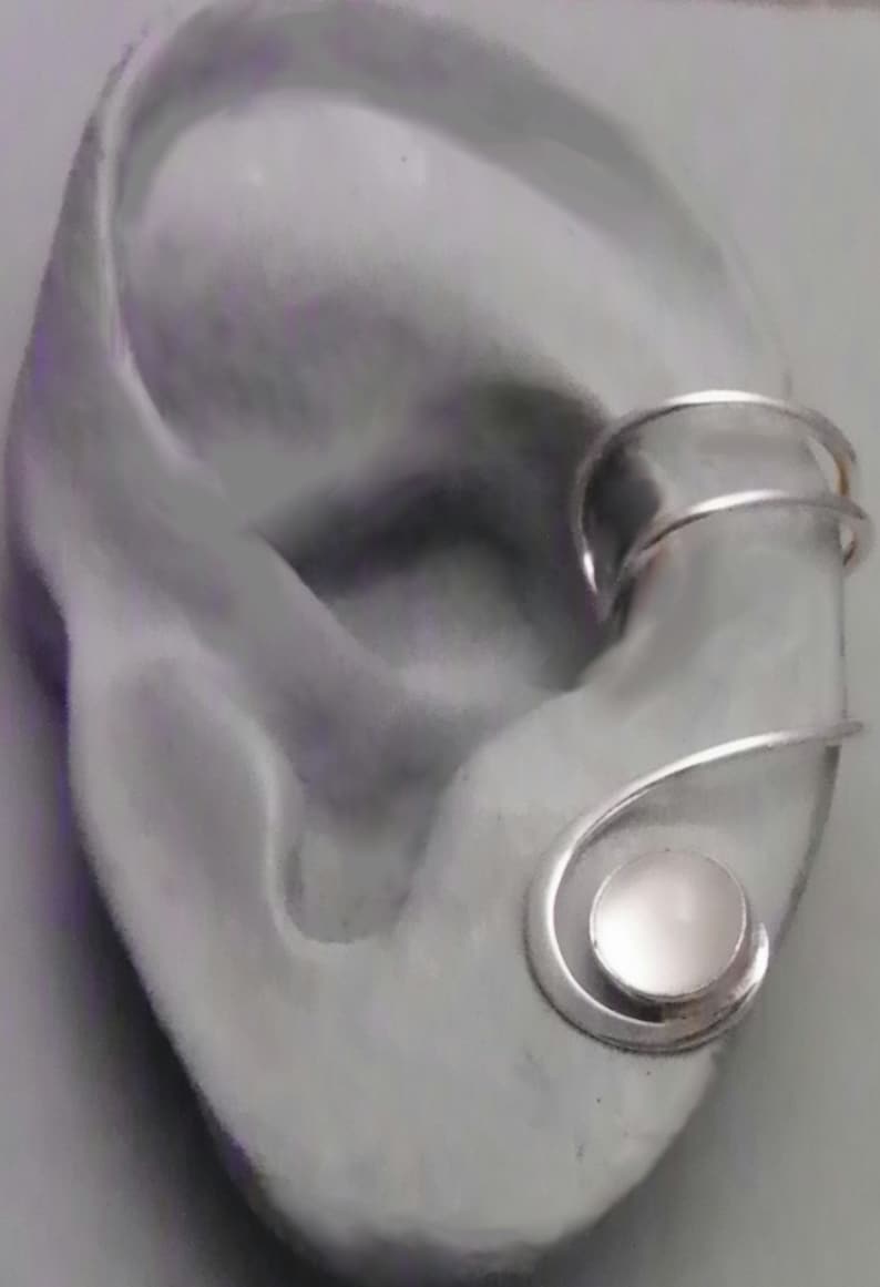 Pode incluir: Um ear cuff de tom prateado com uma banda de fio duplo que envolve a cartilagem superior da orelha e termina numa pedra caboch&atilde;o circular de cor clara, engastada numa estrutura de metal em espiral na parte inferior da orelha.