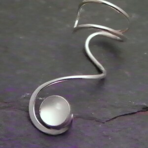 Pode incluir: Um ear cuff de tom prateado com um design de fio cont&iacute;nuo que se curva em forma de espiral. Na extremidade da espiral, uma pequena pedra branca circular e lisa est&aacute; engastada na estrutura met&aacute;lica, criando um aspeto elegante e polido.