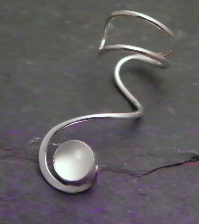 Pode incluir: Um ear cuff de tom prateado com um design de fio cont&iacute;nuo que se curva em forma de espiral. Na extremidade da espiral, uma pequena pedra branca circular e lisa est&aacute; engastada na estrutura met&aacute;lica, criando um aspeto elegante e polido.