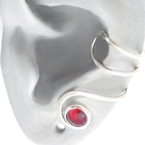 May include: A silver-toned ear cuff featuring a spiral design that wraps around the ear cartilage. The lower end of the metal wire terminates in a circular setting containing a smooth, polished, deep red stone with a swirling pattern.
