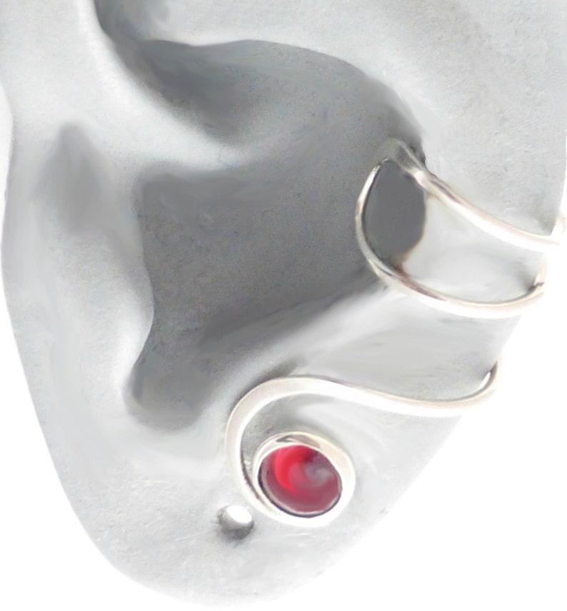 May include: A silver-toned ear cuff featuring a spiral design that wraps around the ear cartilage. The lower end of the metal wire terminates in a circular setting containing a smooth, polished, deep red stone with a swirling pattern.