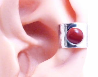 May include: A silver-colored ear cuff featuring a wide, hammered metal band with a smooth, circular, deep red stone set in the center. The metallic surface has an irregular, textured finish that catches the light, contrasting with the stone.