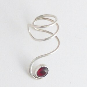 May include: A silver-toned metal ring featuring a continuous, spiraling wire design that wraps around the finger. The end of the wire terminates in a smooth, bezel-set, deep red oval gemstone, creating a fluid and elongated aesthetic shape.