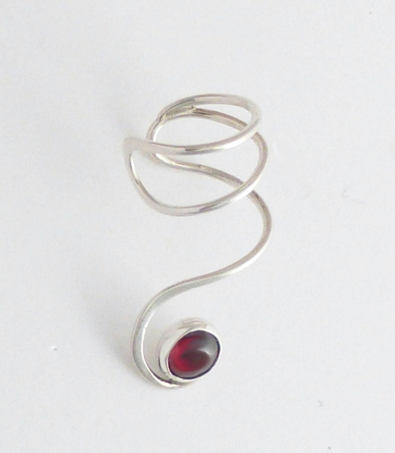 May include: A silver-toned metal ring featuring a continuous, spiraling wire design that wraps around the finger. The end of the wire terminates in a smooth, bezel-set, deep red oval gemstone, creating a fluid and elongated aesthetic shape.