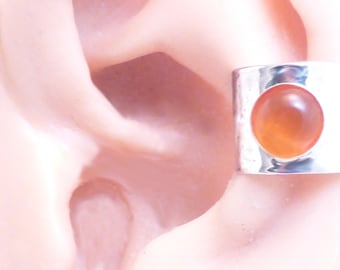 May include: A wide, polished silver ear cuff featuring a single, smooth, circular orange cabochon gemstone set in the center. The metal band has a reflective, metallic finish and wraps around the outer edge of the ear cartilage.