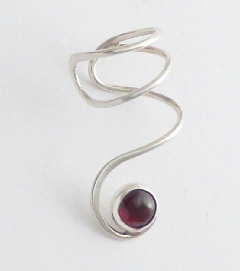 May include: A silver-toned metal ring featuring a spiraling, open-ended band design. The structure curves upward into a circular loop at the top and terminates at the bottom with a single, smooth, dark red cabochon stone set in a silver bezel.