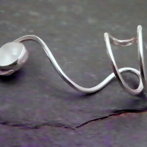 Pode incluir: Um anel de metal prateado com um design curvo e aberto que envolve o dedo. Uma extremidade da banda &eacute; rematada com uma pedra caboch&atilde;o redonda, lisa e branca opaca, enquanto a outra extremidade consiste em dois la&ccedil;os de metal paralelos e curvos.