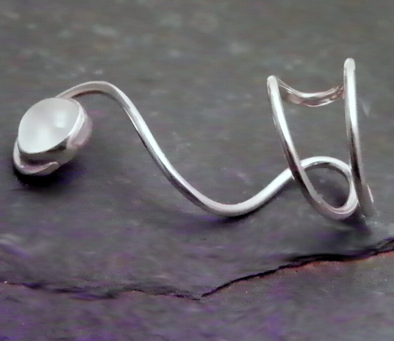 Pode incluir: Um anel de metal prateado com um design curvo e aberto que envolve o dedo. Uma extremidade da banda &eacute; rematada com uma pedra caboch&atilde;o redonda, lisa e branca opaca, enquanto a outra extremidade consiste em dois la&ccedil;os de metal paralelos e curvos.