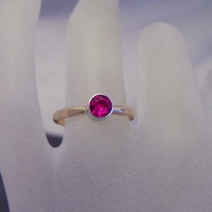 May include: A gold ring with a vibrant, round, magenta-colored gemstone. The gemstone is set in a silver bezel, and the band is a warm gold color. The ring is displayed on a light background.
