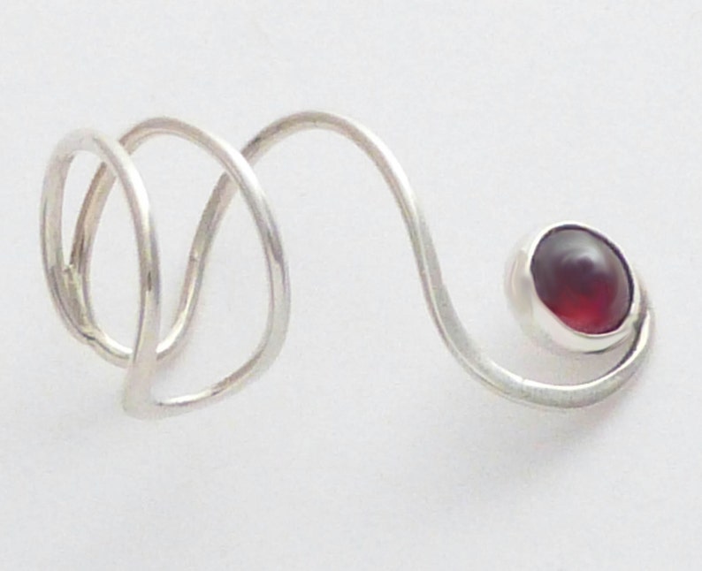 May include: A silver-colored metal ring featuring a spiral design that wraps around the finger. The band extends into a curved arm that terminates in a single, round, deep red gemstone set in a smooth, polished silver-colored metal bezel.