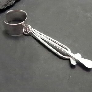 May include: A silver-toned ear cuff featuring a dangling charm composed of three elongated, slender metal rods that are joined at the top and flare out slightly at the bottom, creating a sleek, metallic, and minimalist drop design.